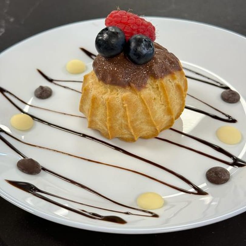 A cream puff with chocolate frosting, blueberries, and a raspberry, on a white plate with chocolate drizzle.
